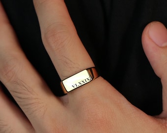 May include: A gold-colored rectangular ring with the inscription "VLXXIX" is worn on a finger. The ring is a simple, modern design, and the hand is against a dark background.