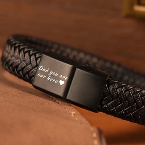 Könnte beinhalten: Schwarzes geflochtenes Lederarmband mit rechteckigem Verschluss. Der Verschluss ist mit den Worten "Dad you are our hero" und einem Herzsymbol graviert. Das Armband wird auf einer braunen Lederoberfläche präsentiert.