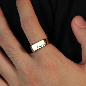 May include: A gold signet ring with the name "Lucas" engraved on the rectangular face. The ring is worn on a finger, showcasing its polished finish and simple design. The ring is a classic piece of jewellery.