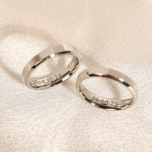 Puede incluir: Dos anillos de boda plateados con fechas grabadas, 09.10.26 y 06.09.25, sobre una superficie de satén crema. Los anillos tienen un diseño plano y facetado. La luz se refleja en el metal.