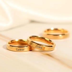 Puede incluir: Tres anillos dorados se muestran sobre una superficie clara. Dos anillos están enfocados, con los nombres "Saber" y "John" grabados. El tercer anillo está desenfocado. Los anillos tienen un diseño sencillo y elegante. El fondo es claro.