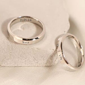 Peut inclure: Deux bagues en argent avec texte gravé. L'une porte l'inscription "01.01.26" et "Rose". L'autre bague porte l'inscription "Lily". Les bagues sont sur une surface claire.