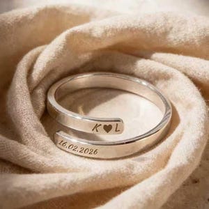 May include: A silver-colored wrap ring with engraved initials "K L" and a heart symbol. The date "16.02.2026" is also engraved on the ring. The ring is displayed on a beige fabric background.