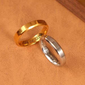Puede incluir: Dos anillos de boda, uno dorado y otro plateado, sobre una superficie marrón. Ambos anillos tienen la fecha "03.02.2025" grabada en el interior. El anillo dorado tiene un acabado pulido, mientras que el plateado tiene un aspecto cepillado.