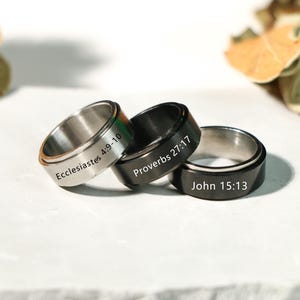 May include: Three spinner rings in silver and black, each engraved with a Bible verse: Ecclesiastes 4:9-10, Proverbs 27:17, and John 15:13. The rings are displayed on a white surface with neutral background elements.