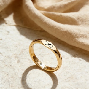 Puede incluir: Un anillo dorado con un símbolo de infinito y la fecha 01.02.2026 grabada. El anillo se muestra sobre una superficie texturizada con una tela beige suave en el fondo. El diseño es simple y elegante.