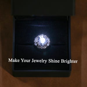 Pu&ograve; includere: Un anello scintillante con una grande pietra centrale e pietre pi&ugrave; piccole intorno, presentato in un cofanetto nero. Il testo "Make Your Jewelry Shine Brighter" &egrave; visibile sotto. L'anello &egrave; illuminato.