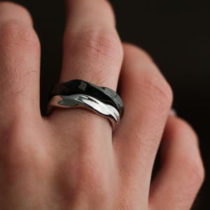 May include: A close-up of two intertwined rings. One ring is silver, and the other is black. The rings have a wavy, textured design. The rings are on a finger, with the hand in the foreground and a dark background.
