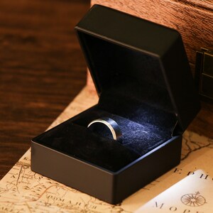 Pu&ograve; includere: Una scatola nera per anelli con un anello d'argento all'interno. L'anello &egrave; centrato nella scatola, foderata in velluto nero. La scatola &egrave; aperta e l'anello &egrave; illuminato da una piccola luce.