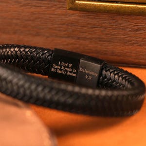 May include: Black braided leather bracelet with a rectangular clasp. The clasp features the text "A Cord Of Three Strands Is Not Easily Broken. Ecclesiastes 4:12". The bracelet is displayed on a wooden surface.