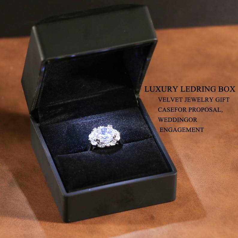 Pu&ograve; includere: Un portagioie nero con interno in velluto, illuminato per mostrare un anello di diamanti. L'anello presenta una grande pietra rotonda e trasparente circondata da pietre pi&ugrave; piccole. Testo: LUXURY LEDRING BOX, VELVET JEWELRY GIFT CASEFOR PROPOSAL, WEDDINGOR ENGAGEMENT.