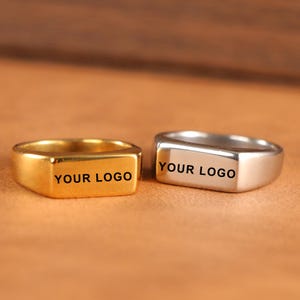以下が含まれることがあります： 長方形の面を持つ2つのシグネットリング。1つは金色、もう1つは銀色です。「YOUR LOGO」の文字が各リングに黒で印刷されています。リングは茶色の表面に表示されています。