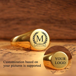 May include: A gold signet ring with a circular face. The face features a black letter "M" within a laurel wreath, the name "MARSON", and the date "08.06.2025". The ring is displayed on a textured surface. The text "Customization based on your pictures is supported" is also visible.