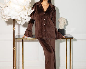 May include: A dark brown velvet pajama set consisting of a long-sleeved button-down shirt with a collar and ruffled cuffs, paired with matching wide-leg trousers featuring ruffled hems. The fabric has a soft, plush texture throughout.