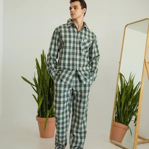 May include: A green, white, and tan plaid pajama set. The long-sleeved button-down shirt has a chest pocket. The matching pants have a relaxed fit. The person is standing barefoot in front of a mirror and potted plants.