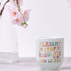 May include: A white ceramic mug featuring a colorful graphic print with the text ALRIGHT STOP REGULATE AND LISTEN. The design includes retro-style lettering in muted tones of yellow, pink, orange, and blue, accented with small flower and smiley face icons.