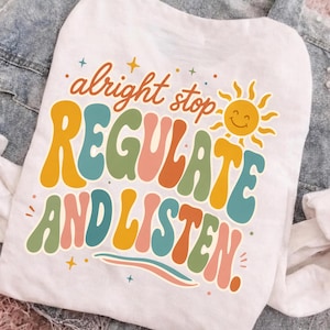 Puede incluir: Una camiseta blanca con un gráfico colorido y el texto "alright stop REGULATE AND LISTEN." El diseño incluye un icono de sol amarillo y pequeños detalles de estrellas en una fuente retro con letras onduladas y multicolores en amarillo, verde azulado, verde y naranja.