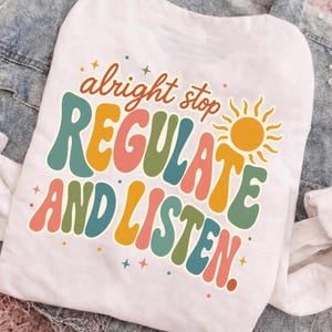 May include: A white t-shirt featuring a colorful graphic print with the text "alright stop REGULATE AND LISTEN." The design uses a retro-inspired font in shades of teal, mustard yellow, and coral, accented by a yellow sun icon and small stars.