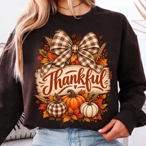 Op de afbeelding: Een donkerbruin sweatshirt met lange mouwen en een grafische print van een grote bruin-witte geruite strik boven een houten bord met het woord Thankful in oranje schrift, omringd door oranje pompoenen en herfstbladeren.