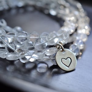 Op de afbeelding: Een armband van heldere kwarts kralen met een zilveren hartbedel. De armband bestaat uit meerdere strengen heldere, afgeronde kralen. De bedel is een kleine, zilveren schijf met een uitgesneden hart.