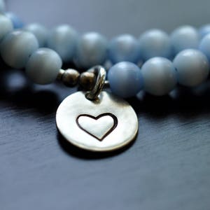 Op de afbeelding: Een zilveren armband met een hartvormige bedel. De armband is gemaakt van lichtblauwe kralen.