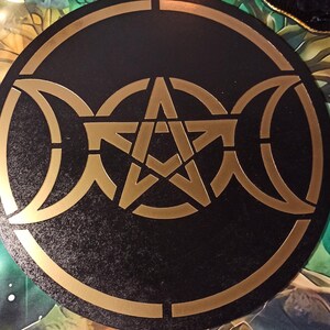 Puede incluir: Un tablero circular de madera negro con un pentagrama dorado y un símbolo de triple luna. El tablero está rodeado por un borde dorado.