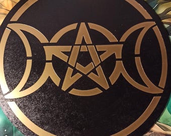 May include: A black circular wooden board with a gold pentagram and triple moon symbol. The board is surrounded by a gold border.