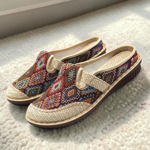 May include: A pair of slip-on shoes featuring a woven fabric upper with a multi-coloured geometric diamond pattern in shades of red, blue, and brown. The shoes have a beige textured trim, a light-coloured interior lining, and a dark brown sole.
