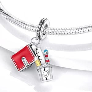 May include: A silver charm with a red book, ruler, pencil, and paintbrushes. The charm is attached to a silver bracelet. The book has white stitching and a rhinestone accent. The pencil is yellow, and the paintbrushes are blue and red.
