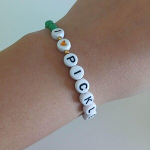 May include: A green and white beaded bracelet with the words "I love pickles" spelled out in black letters.