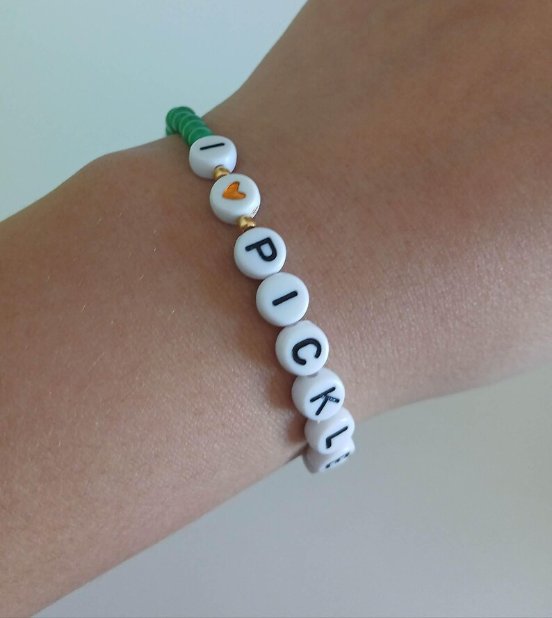 May include: A green and white beaded bracelet with the words "I love pickles" spelled out in black letters.