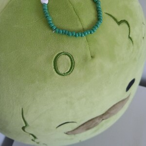 May include: A green plush toy shaped like a pickle with a brown mustache. The toy is wearing a green beaded bracelet with white letter beads that spell "I LOVE PICKLES".