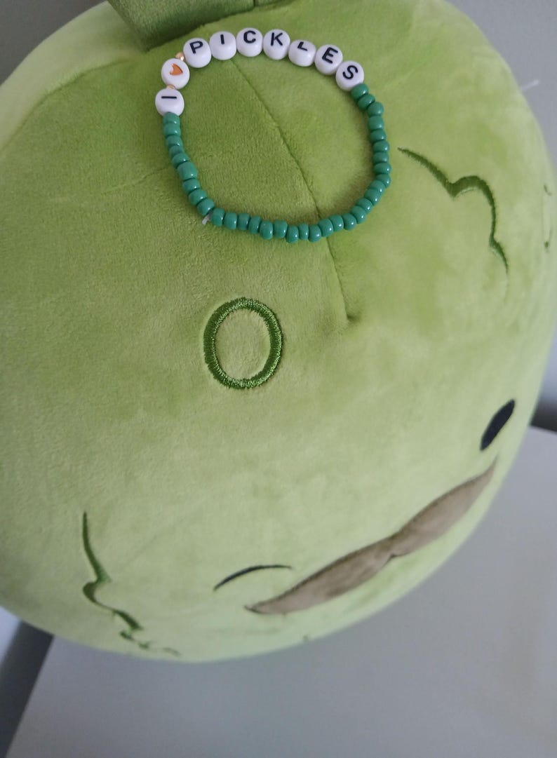 May include: A green plush toy shaped like a pickle with a brown mustache. The toy is wearing a green beaded bracelet with white letter beads that spell "I LOVE PICKLES".