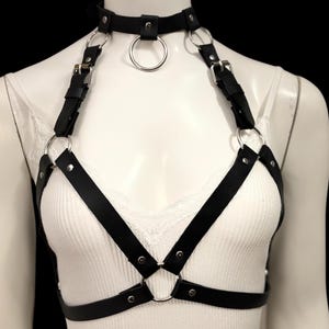 May include: Black leather body harness with silver-tone metal rings and buckles. The harness features a choker-style collar with a large ring, straps over the shoulders, and a series of connecting straps across the chest and torso.