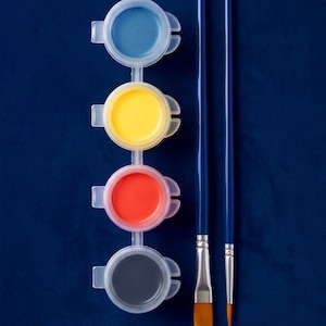 May include: A strip of four connected translucent plastic paint pots containing blue, yellow, red, and dark gray paint, positioned vertically next to two paintbrushes with long blue handles, one with a flat tip and one with a pointed tip.