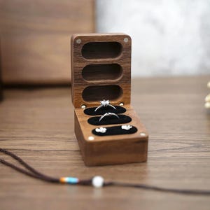 May include: A wooden ring box with three compartments, each holding a ring. The box is open, revealing the rings on a black velvet lining. The box is made of dark wood and has a magnetic closure.