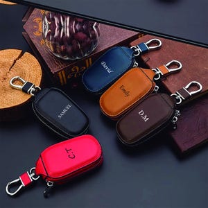 May include: A collection of personalized leather keychains in various colors, including black, blue, brown, tan, and red. Each keychain has a silver-tone clasp and zipper, with names or initials engraved on the front. The keychains are arranged on a dark surface.