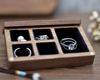 May include: A wooden jewelry box with a hinged lid, featuring six compartments lined with black velvet. The box contains various pieces of jewelry, including rings and a pearl. The box is made of dark wood and sits on a wooden surface.