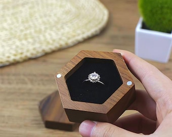 Peut inclure: Une bague de fiançailles en argent avec un halo de petites pierres est présentée dans un écrin hexagonal en bois. La boîte est ouverte, révélant la bague sur un intérieur en velours noir. La boîte est marron avec une fermeture magnétique.
