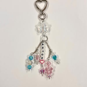 May include: A beaded keychain with a silver heart-shaped clasp. The keychain features the name "ANTON" spelled out in white letter beads. It includes clear star-shaped beads, pink and blue beads, and heart-shaped beads.