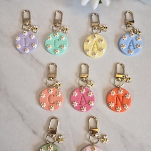 May include: A collection of personalized keychains with gold-colored hardware. Each round charm features a different pastel color, a letter, and small daisy accents. A small bee charm hangs from each keychain.