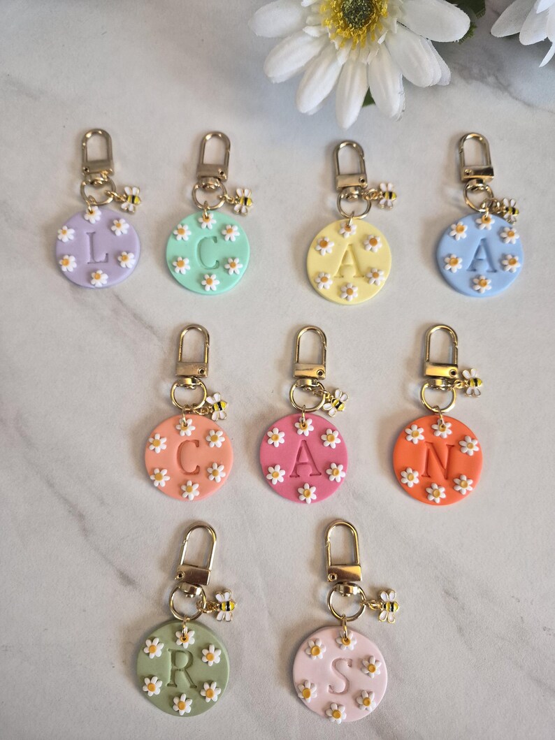 May include: A collection of personalized keychains with gold-colored hardware. Each round charm features a different pastel color, a letter, and small daisy accents. A small bee charm hangs from each keychain.