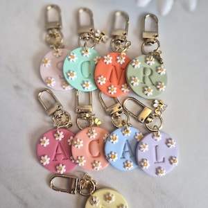 May include: A collection of colorful, daisy-adorned keychains. Each round charm features a different initial letter and is attached to a gold-colored clasp. The keychains are in various pastel colors, including pink, orange, green, and blue.
