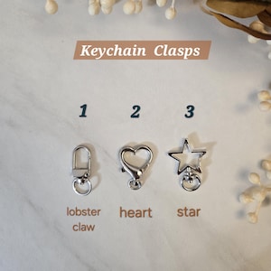 May include: Image of three silver keychain clasps: a lobster claw, a heart, and a star. The text "Keychain Clasps" is displayed above the clasps. The clasps are on a white marble surface with decorative floral elements.
