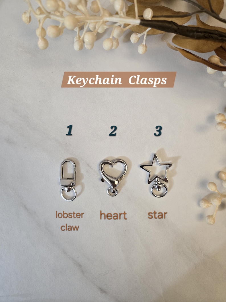 May include: Image of three silver keychain clasps: a lobster claw, a heart, and a star. The text "Keychain Clasps" is displayed above the clasps. The clasps are on a white marble surface with decorative floral elements.