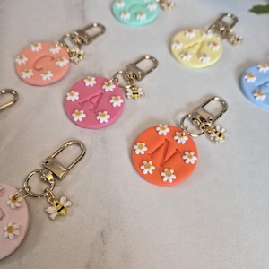 May include: A collection of colorful, round keychains. Each keychain features a different initial letter and is decorated with small white daisies. A gold-colored bee charm hangs from each keychain.