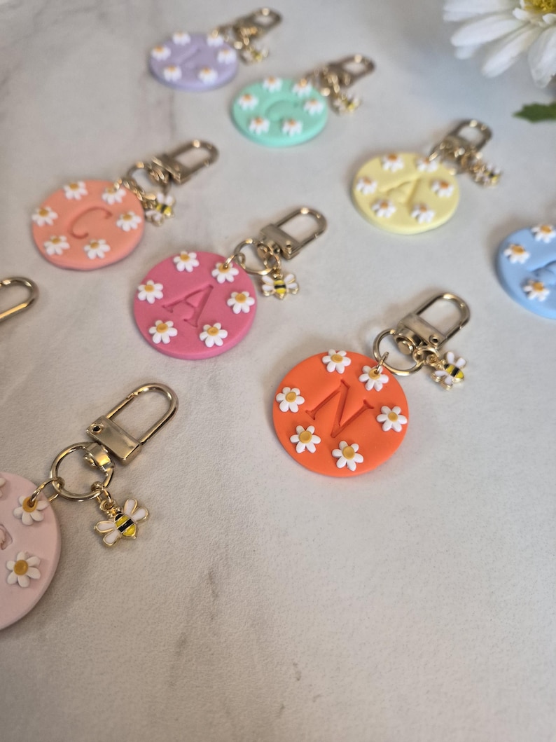 May include: A collection of colorful, round keychains. Each keychain features a different initial letter and is decorated with small white daisies. A gold-colored bee charm hangs from each keychain.