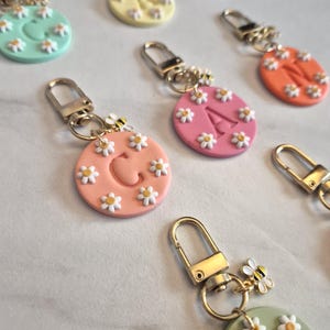 May include: A collection of colorful keychains with daisy and bee accents. Each keychain features a round, pastel-colored base with a letter or shape, surrounded by small white daisies. Gold-colored hardware completes the design.
