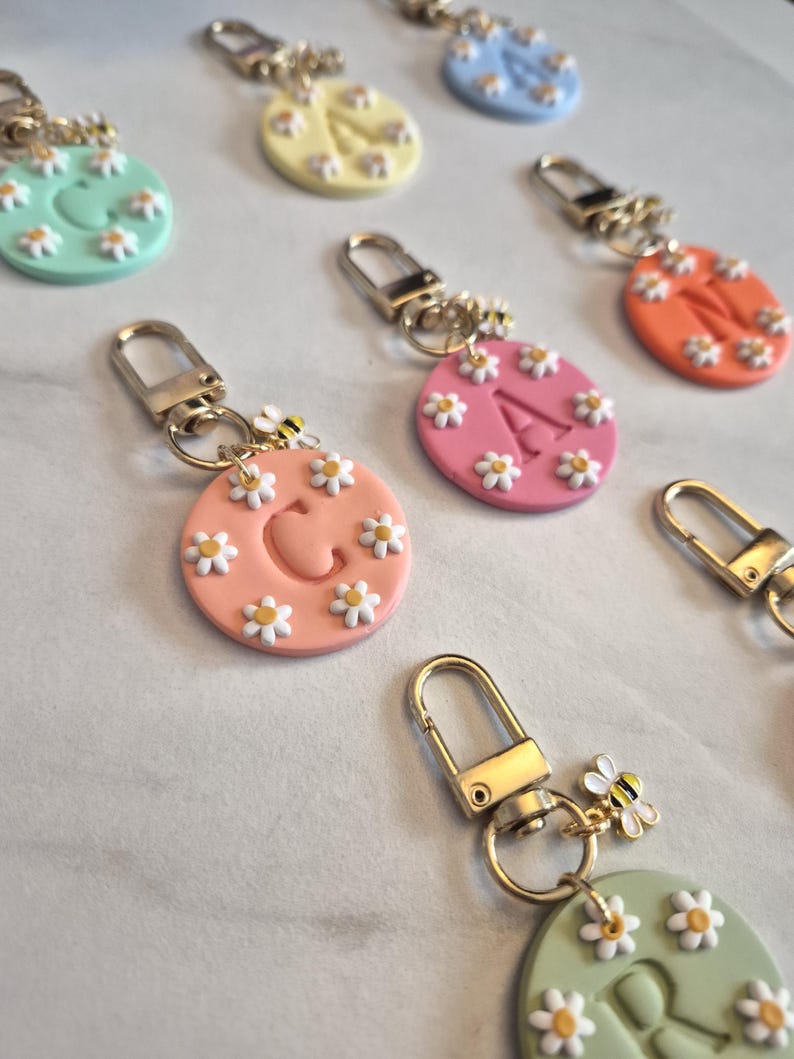 May include: A collection of colorful keychains with daisy and bee accents. Each keychain features a round, pastel-colored base with a letter or shape, surrounded by small white daisies. Gold-colored hardware completes the design.