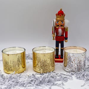 May include: Three scented candles in gold and silver mercury glass containers with white wax. A decorative wooden nutcracker soldier stands behind the candles. The candles are on a white surface with silver paper.